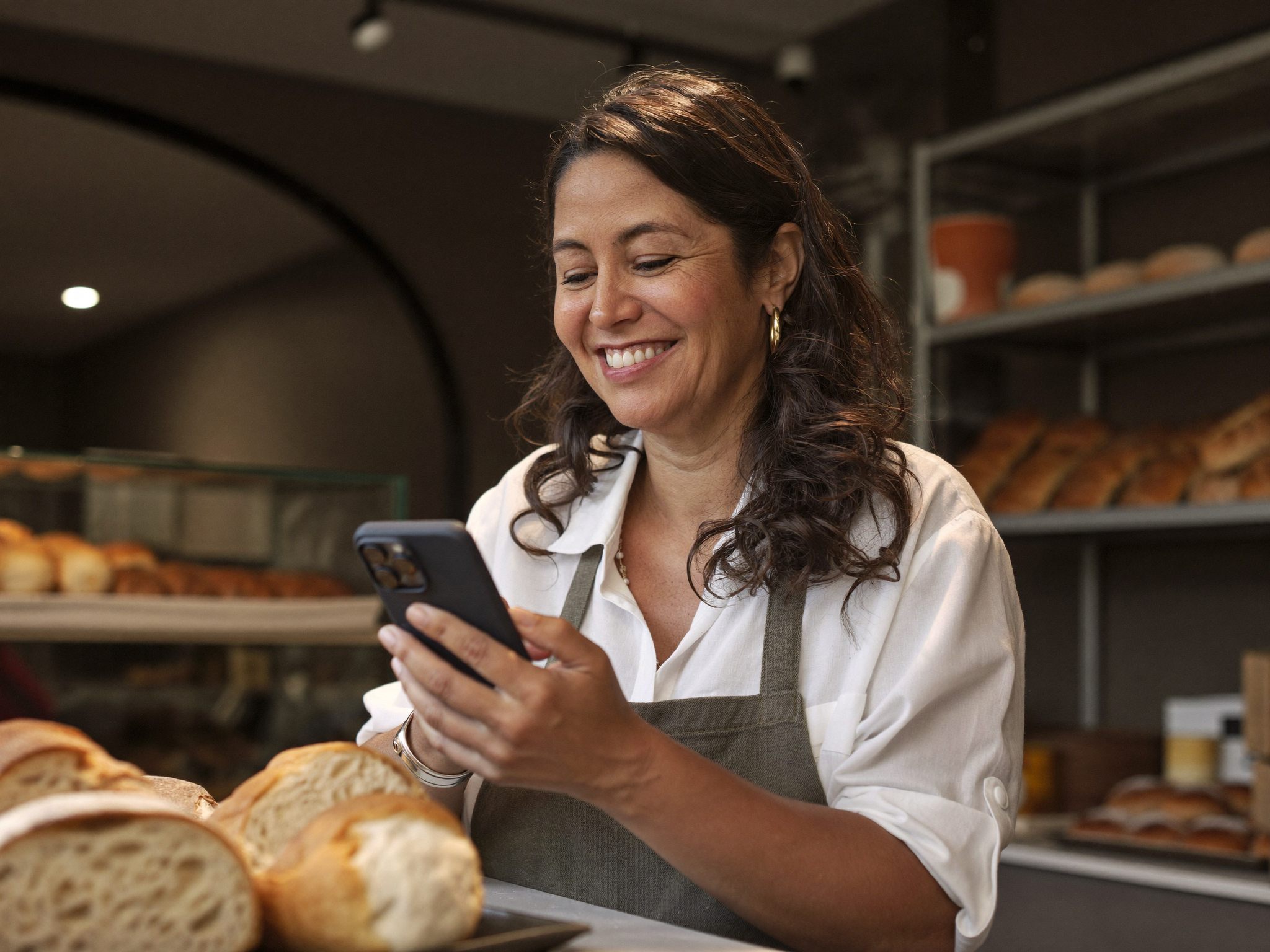 Empreendedora sorrindo ao ver uma mensagem no celular dentro de sua padaria
